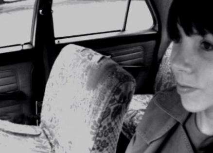 Katrīna Neiburga sēž mašīnā pie autovadītāja stūres. 
